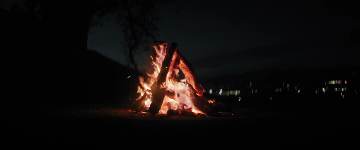 Bonfire burns in a field with the city lights in the background at night