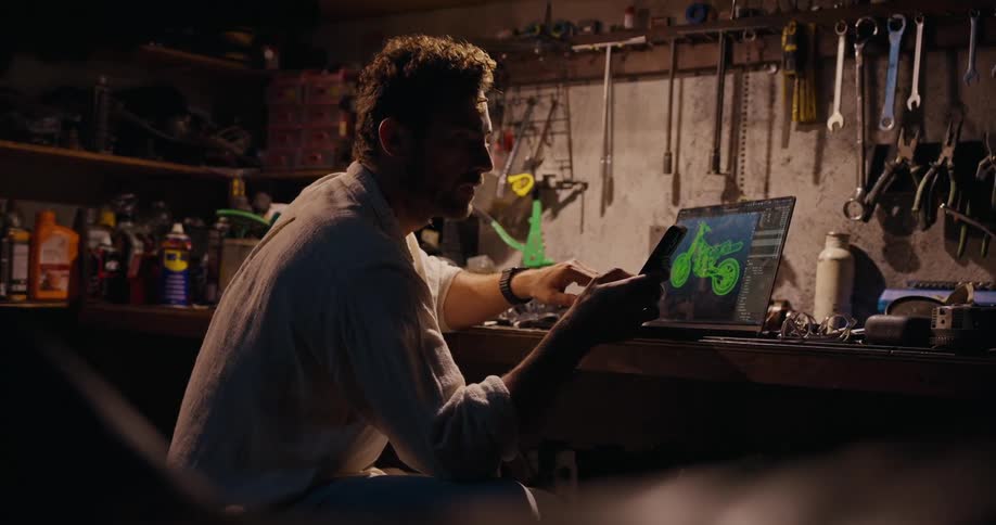 Man inside a workshop typing on a smartphone and next to him tools on a laptop