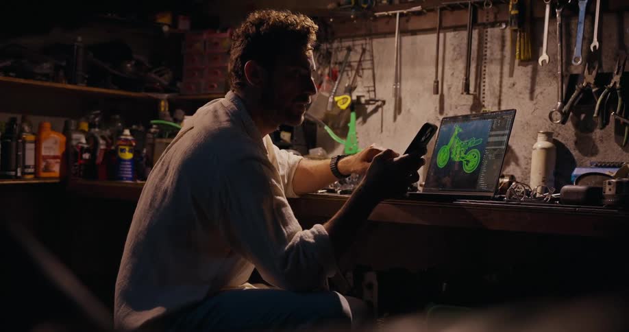A man in a shirt sits at a table using a smartphone and working on a laptop with a 3d motorcycle model