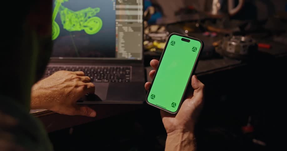 A person sits at a table holding a smartphone with a green screen and working on a laptop with a 3d motorcycle model visible