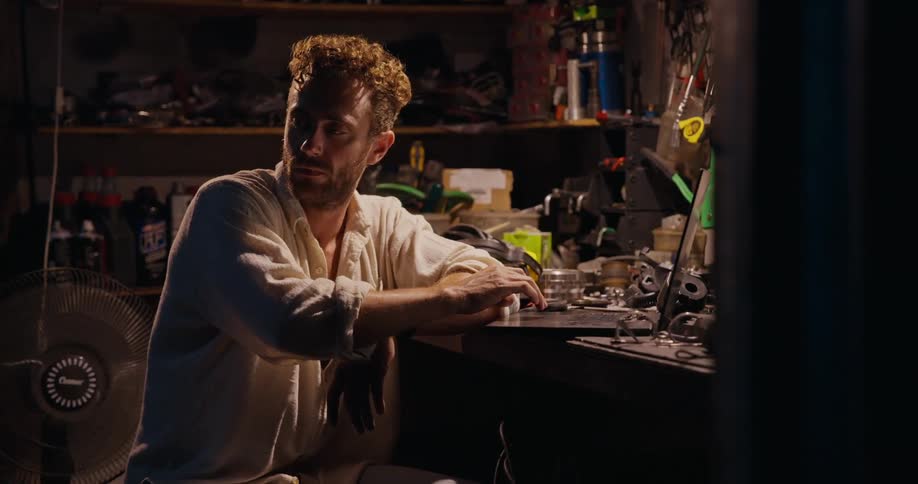 A person at a cluttered workbench in a dimly lit workshop surrounded by tools and mechanical parts