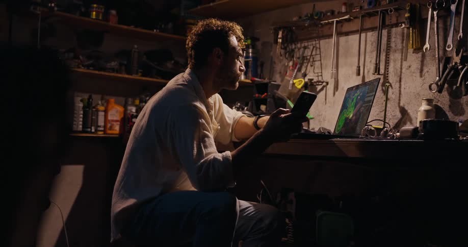 The image shows a person at a cluttered workbench in a dimly lit workshop surrounded by tools and mechanical parts