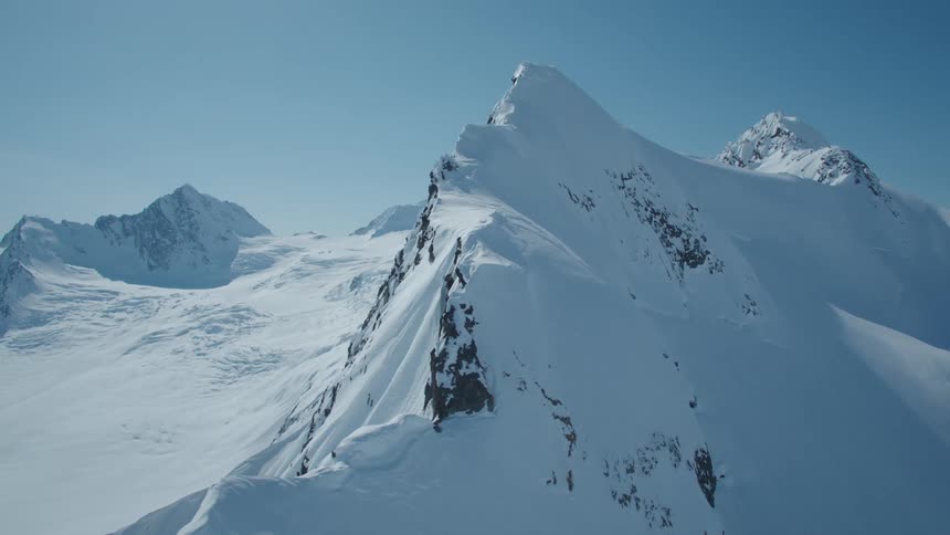 Aerial view showcases a majestic mountain range blanketed in snow with a sharp peak dominating the scene under a clear blue sky