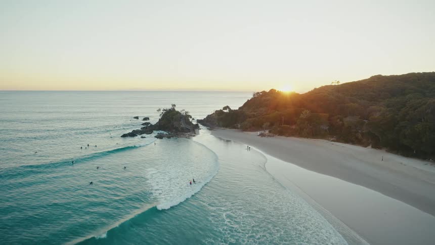 People surf and swim in the ocean near a beach as the sun hangs low in the sky