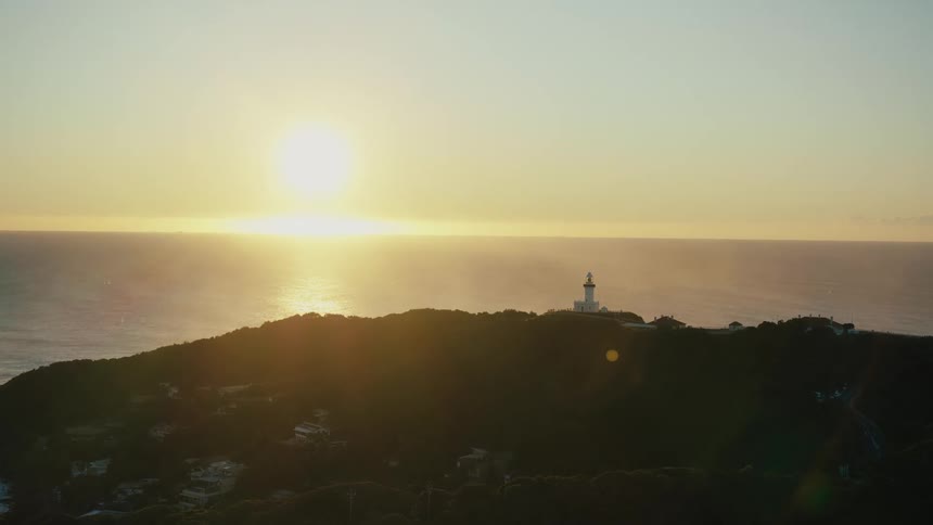 A lighthouse sits atop a cliff overlooking the sun setting over the ocean