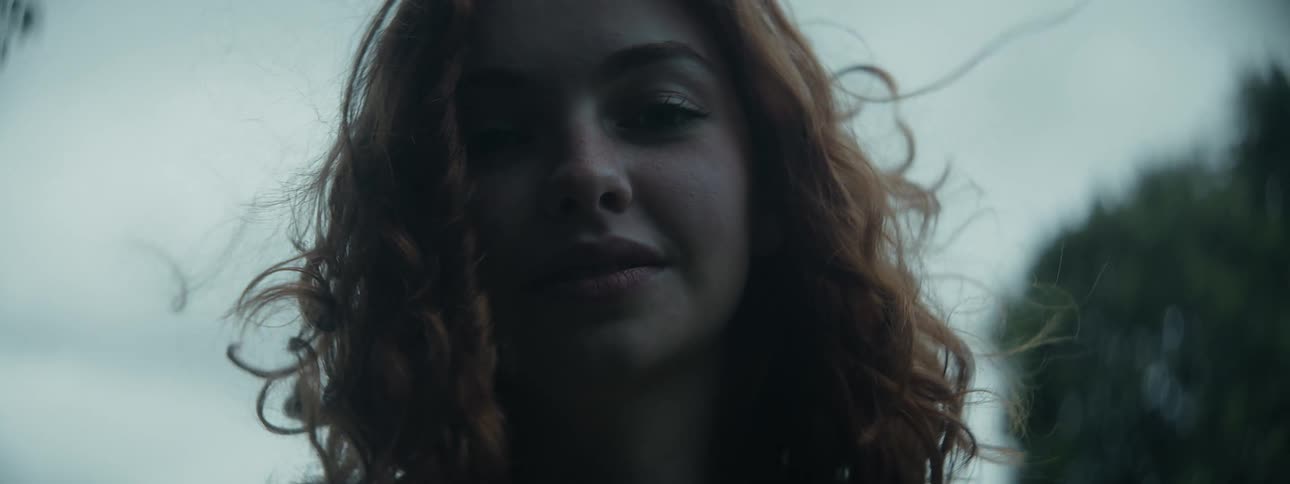A smiling teenage girl with red hair and wearing a sweater stands outside as the wind blows her hair