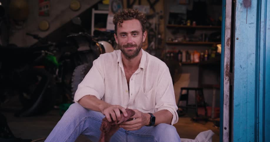 A person sits on the floor of a garage holding a cloth there is a motorcycle in the background and various tools on shelves