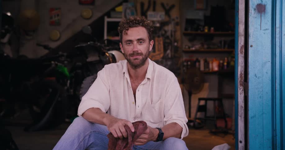 A person sits in a garage holding a cloth with tools and a motorcycle in the background