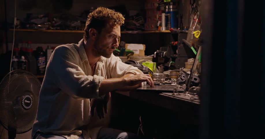 A person at a cluttered workbench in a dimly lit workshop surrounded by tools and mechanical parts