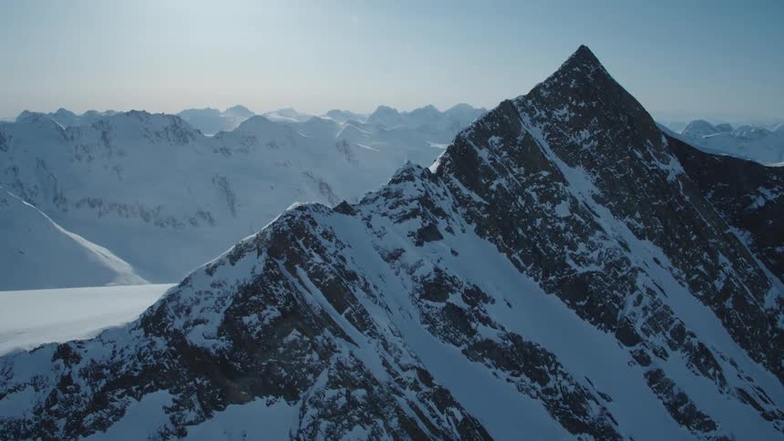An aerial view showcases a majestic mountain range blanketed in snow with a sharp peak dominating the scene under a clear blue sky