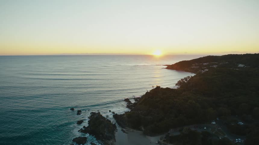 Buildings dot a coast with waves rolling into a beach as the sun sits on the horizon