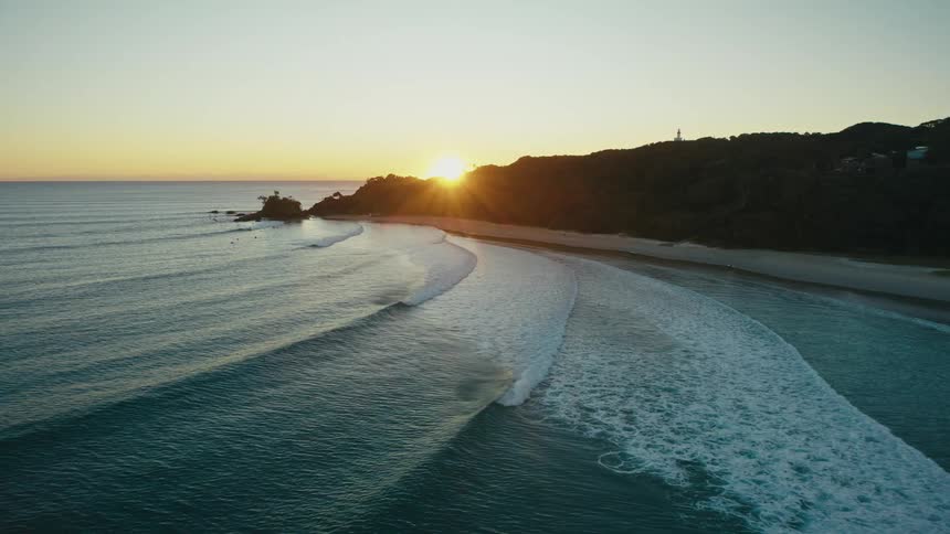 A serene coastal scene at twilight with rolling waves gently lapping against a sandy beach backed by a verdant hillside dotted with houses and a lighthouse