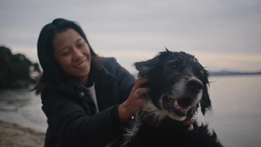 A woman pets a wet dog on a beach with a lake and a cloudy sky in the background