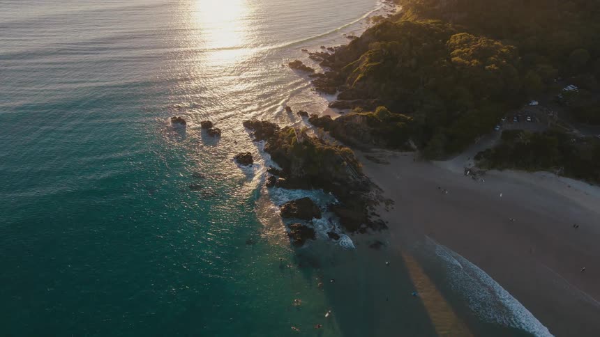 Aerial view of a sundrenched coastline where golden rays dance on the turquoise water highlighting rocky outcrops and a pristine sandy beach