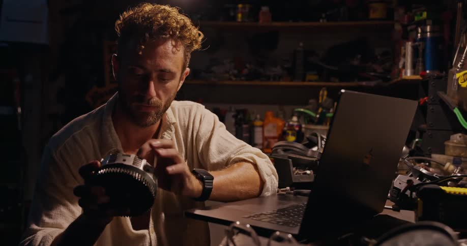 Mechanic in a workshop working on a laptop and picking up a cylindrical part from a motorcycle