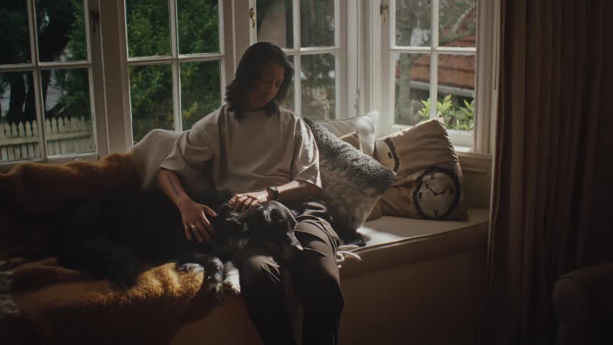 A woman sits on a couch at home petting a dog near a window