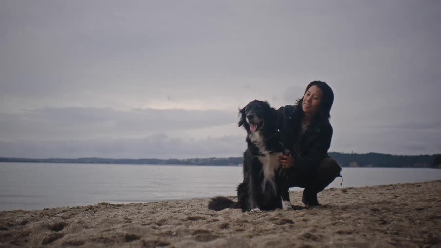 A young asian man kneels on the beach while petting his black dog