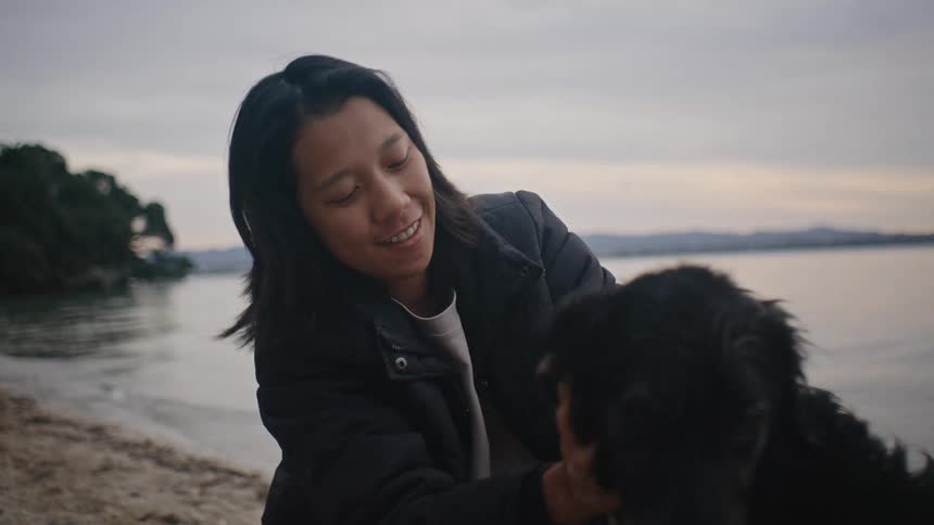 An asian woman kneels on a beach and smiles as she pets a black dog