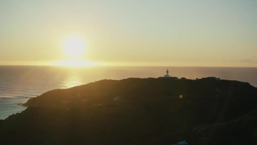 Sun tints the sky and the ocean waters between the horizon and the lighthouse topped shore red