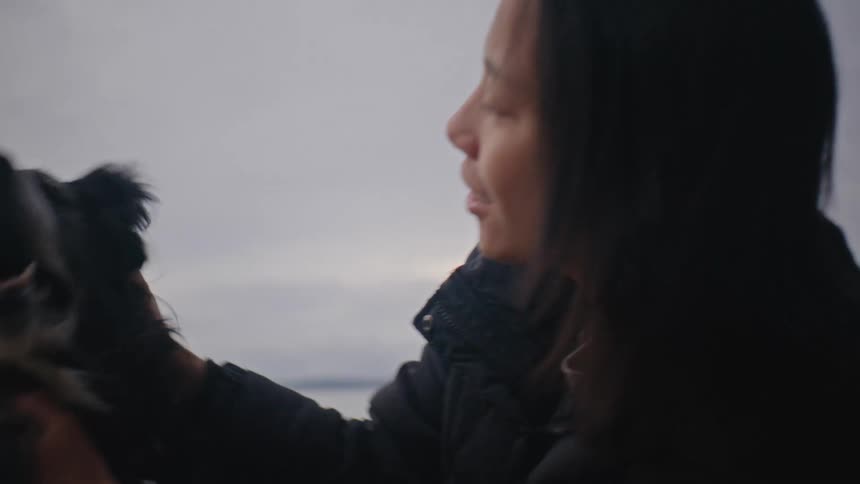 A woman in a black coat pets a black and white dog on a cloudy beach
