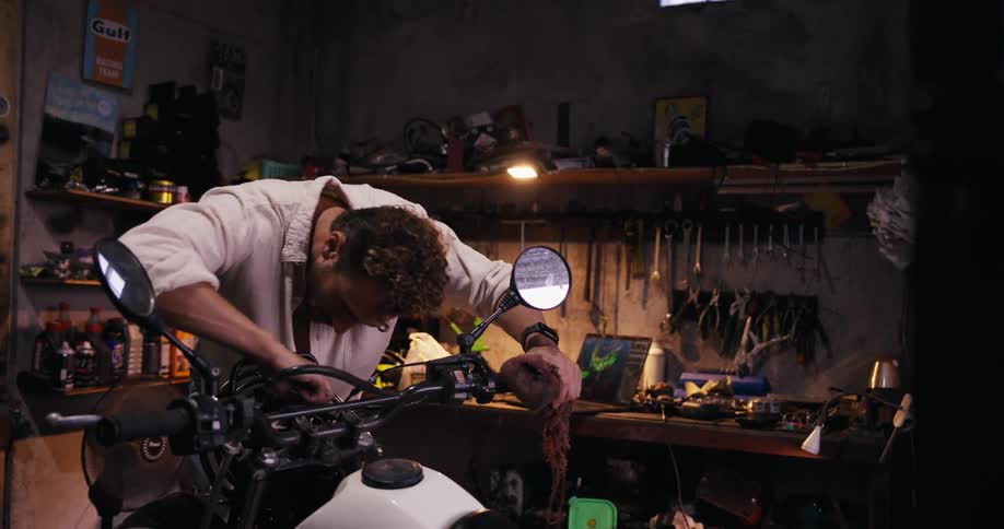 A man is working intensely on a motorcycle in a cluttered garage surrounded by tools and mechanical parts with dirt on his hands and a focused expression