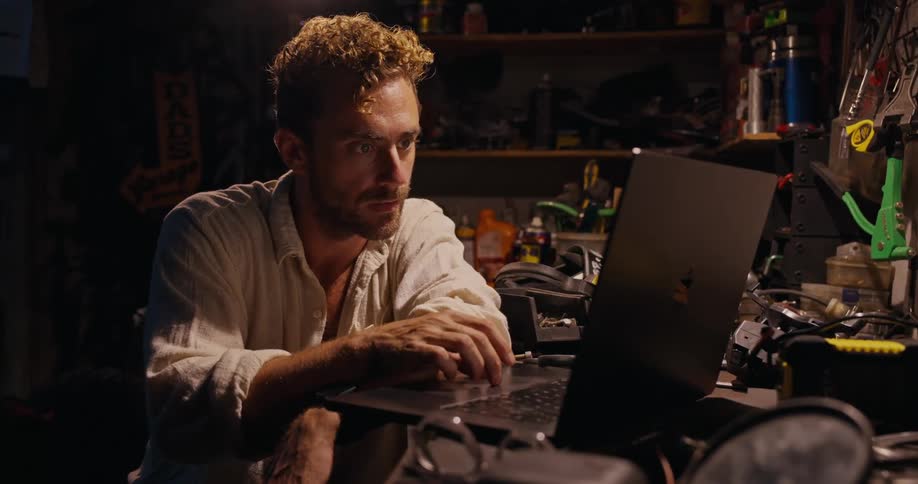 A man in a white shirt sits in a dimly lit workspace and uses a laptop