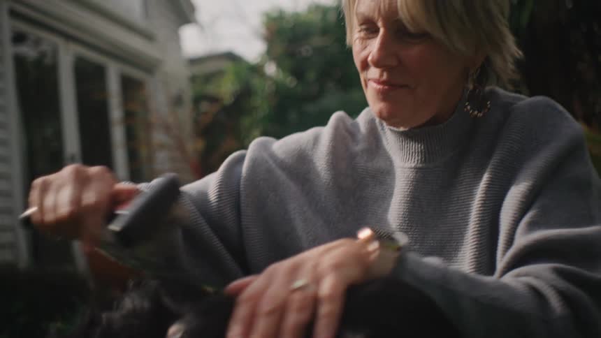 A woman with gray hair wearing hoop earrings a sweater and a gold watch pets and brushes her dog