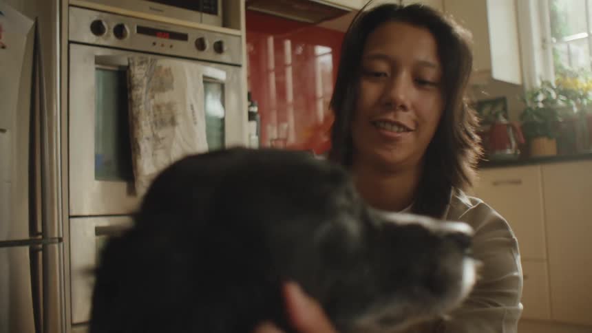 A woman with straight hair and a gray shirt pets a black dog near a kitchen oven