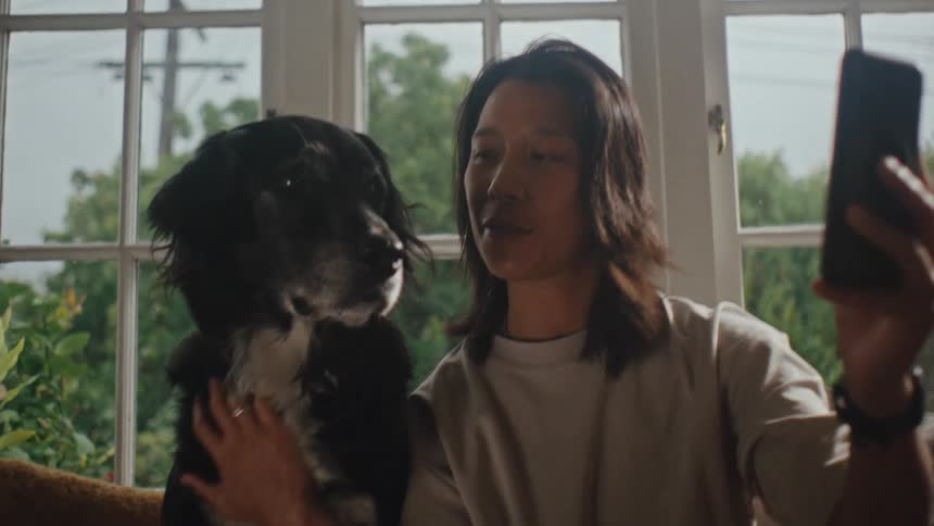 Man sitting in his living room smiles and pets his dog while taking a selfie