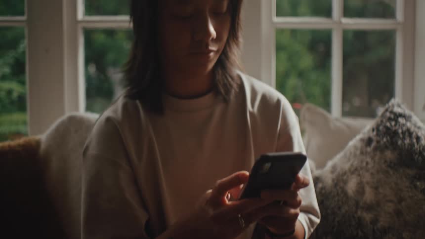 A person sits on a couch holding a smartphone next to a window