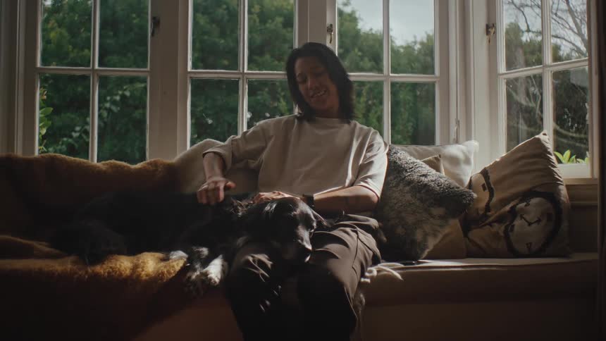 A woman sits on a couch petting a black dog that is resting on her lap in the living room smiling
