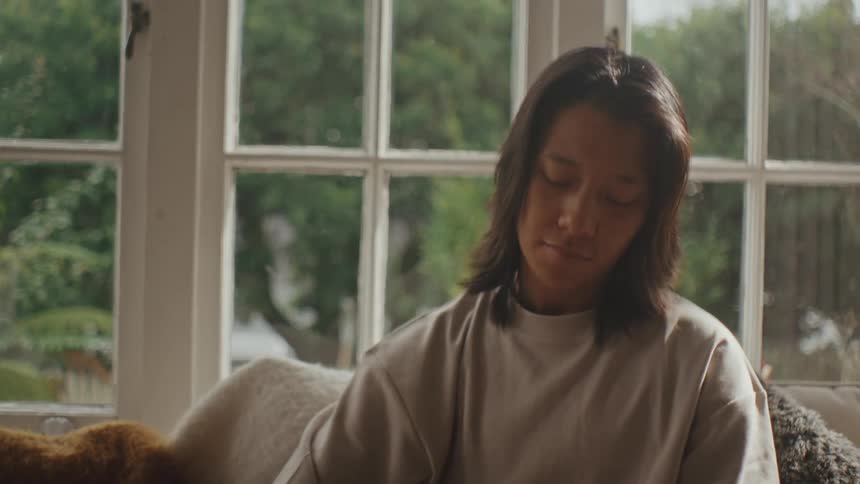 A woman with straight hair and a gray shirt sits on a couch petting a black dog that is resting on their lap