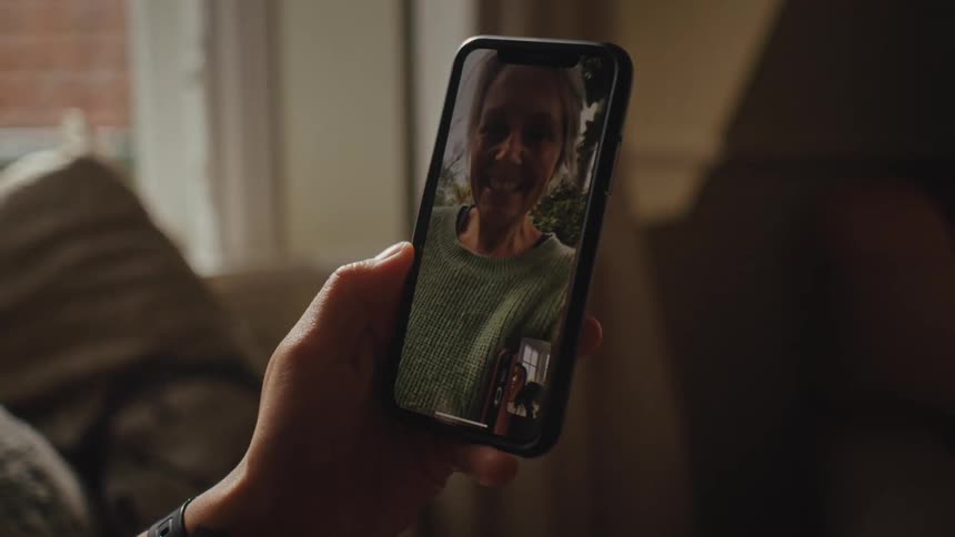 A hand holds a smartphone displaying a video call with another person in a cozy indoor setting with a couch and window in the background