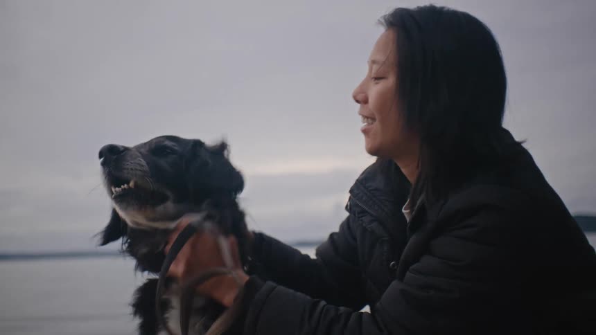 Woman wearing a black coat strokes a black and white dog on the beach on a cloudy day