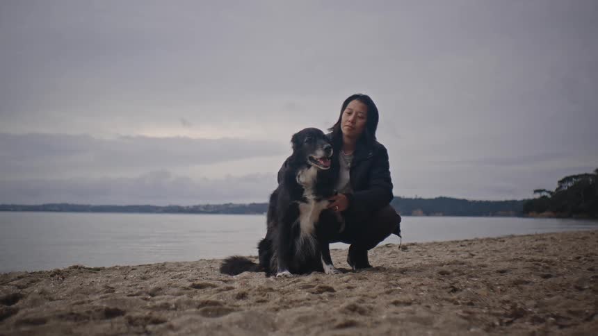 Woman wearing a black jacket pets a black and white dog on the beach near the sea