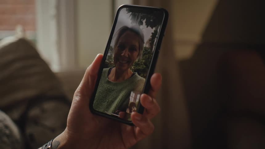 A hand holds a smartphone displaying a video call with another person in a cozy indoor setting with a couch and window in the background