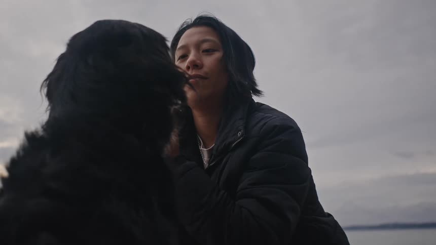 A woman pets a dog on a beach with a lake and a cloudy sky in the background