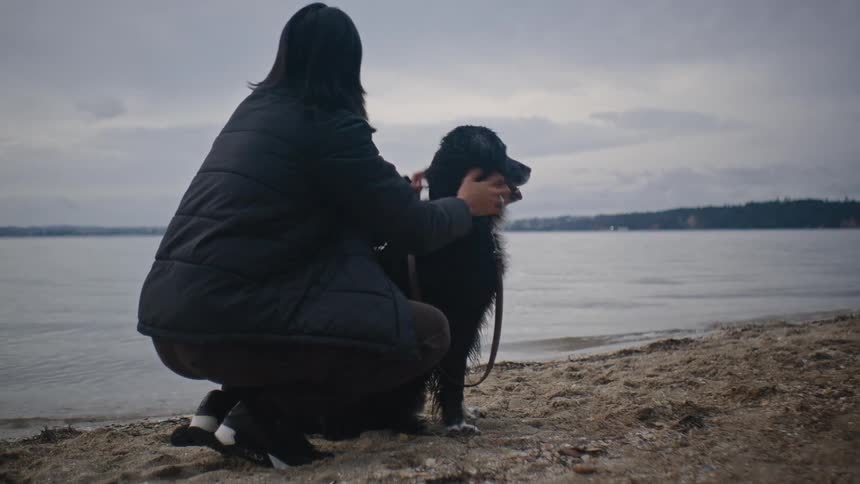 Woman in a black coat strokes a black and white dog on the beach near the sea