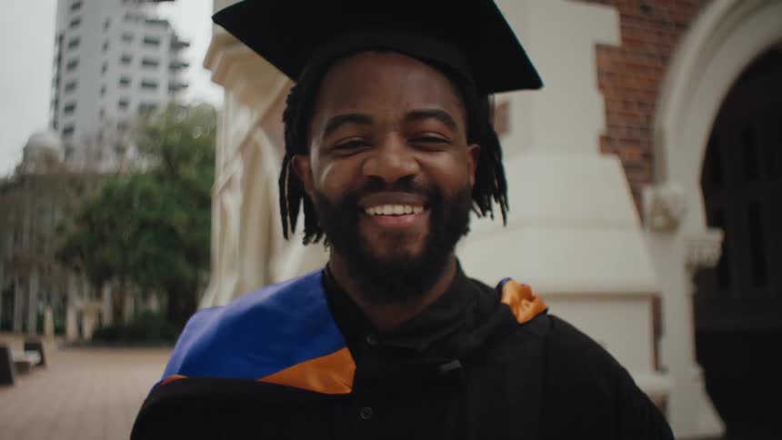 A student in a cap and gown smiles brightly in front of a brick building