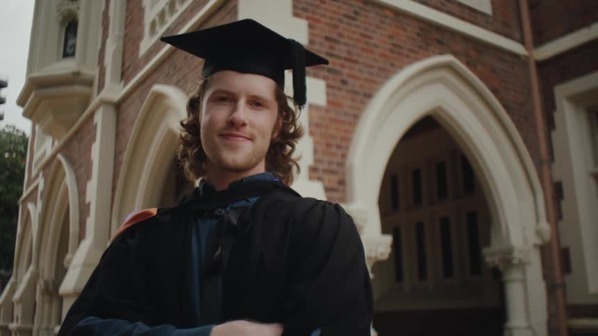 A person wearing a graduation cap and gown stands in front of a brick building with arched doorways
