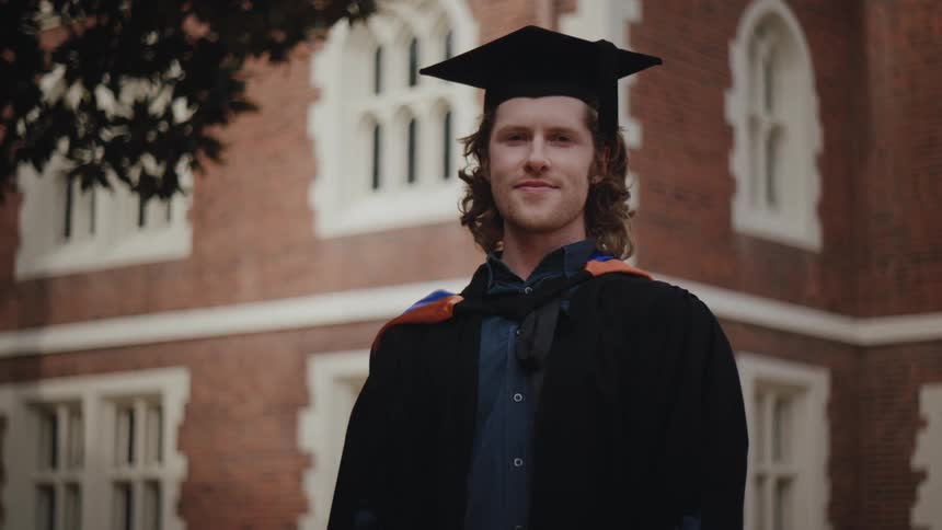 A young man in front of a school building outdoors in a graduation cap and gown smiles at the camera