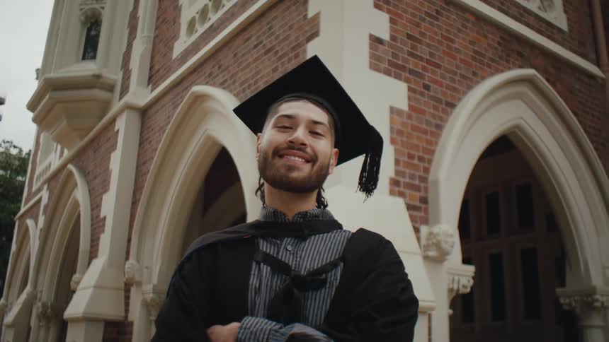 A man is wearing college graduation attire smiling at the camera which zooms in on him