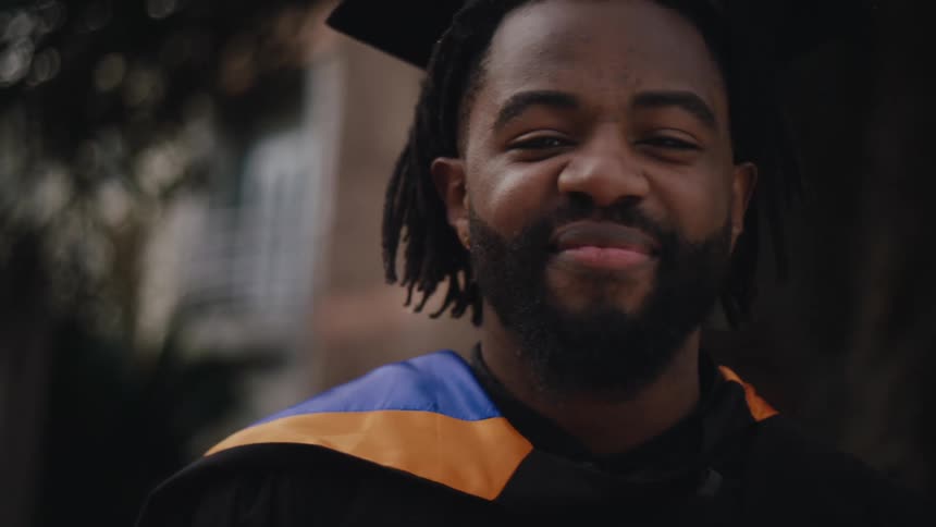 A joyful graduate with dreadlocks wears blue and gold academic robes