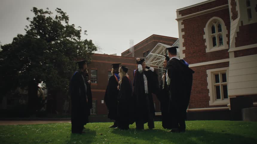 A group of students who just graduated from school stand around in their robes and mortarboard hats talking outside of the school