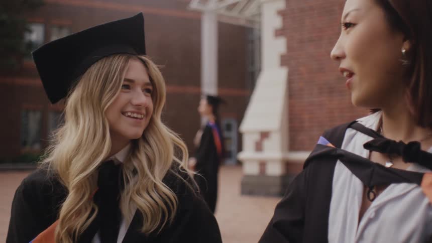 Two friends wearing graduation gowns happily talking to each other