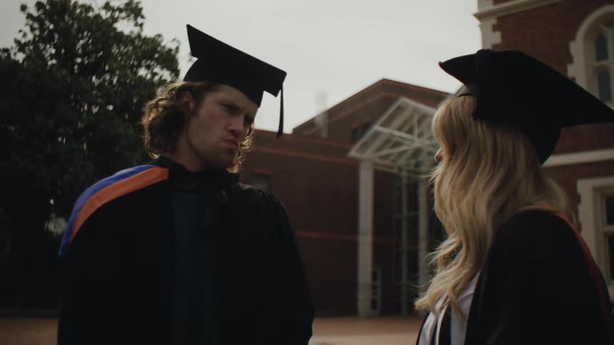 Two students dressed in graduation attire have a conversation outside of a school building