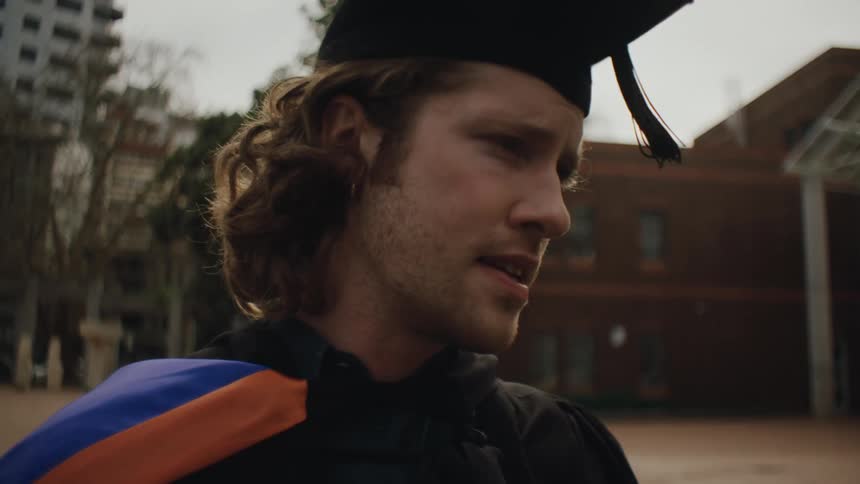 Man wearing graduation cap speaks while standing in front of a building outdoors