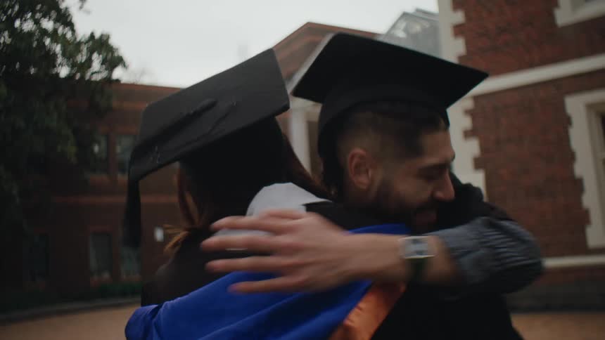 Men wearing graduation cap and gown embraces friend outdoors