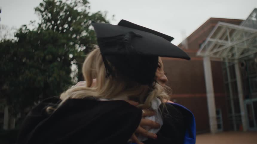 Two graduate students in gowns and caps hug each other in celebration