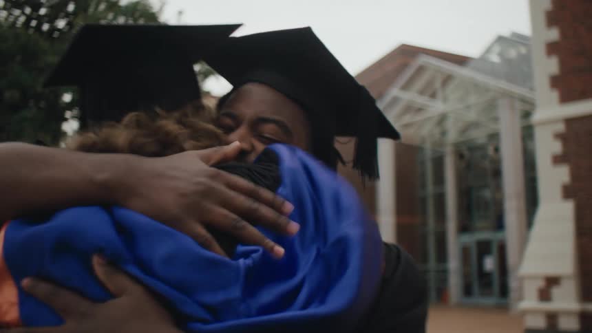 Graduating seniors wearing their graduation robes and caps hug while standing near buildings on campus